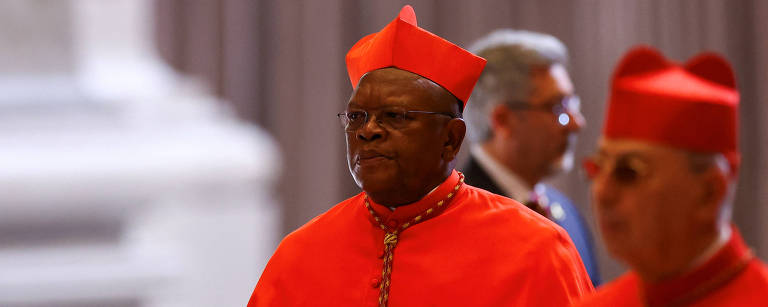 A imagem mostra dois cardeais vestidos com vestes litúrgicas vermelhas. O cardeal à esquerda está segurando papéis e parece estar em um momento de reflexão. O cardeal à direita também está vestido de vermelho, mas com um detalhe em dourado. Ao fundo, é possível ver outras pessoas e a arquitetura de um espaço religioso.