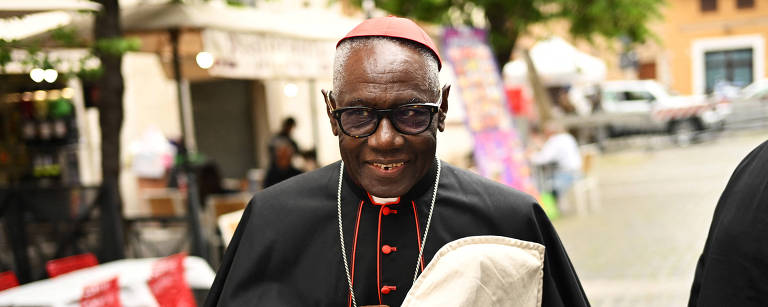 Um cardeal está sorrindo e segurando um objeto em suas mãos. Ele usa vestes religiosas tradicionais, predominantemente pretas com detalhes vermelhos. O fundo mostra mesas e cadeiras vermelhas em um ambiente urbano, com algumas pessoas visíveis ao fundo. O cardeal usa óculos e tem uma expressão amigável.