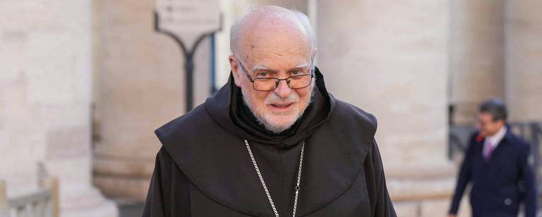 Um homem idoso, vestido com um hábito religioso escuro e um cordão com um crucifixo, caminha em um ambiente externo. Ele usa óculos e tem cabelo grisalho. Ao fundo, há colunas e outras pessoas passando.