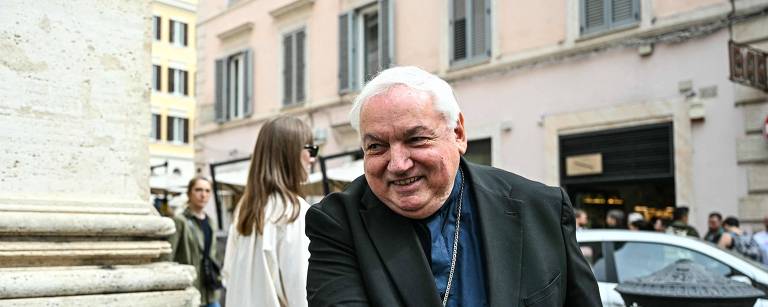 Um homem sorridente, vestido com um traje escuro, está cumprimentando outra pessoa com um aperto de mão. Ao fundo, há pessoas caminhando na rua, algumas delas vestindo roupas casuais. O cenário é urbano, com edifícios e lojas visíveis. O homem parece estar em um momento amigável e descontraído.