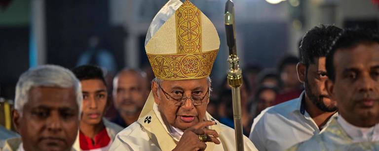 A imagem mostra um grupo de pessoas em uma cerimônia religiosa. No centro, um homem idoso usa um manto branco e um chapéu dourado, segurando um cajado. Ele é cercado por outros homens vestidos com vestes religiosas claras. Ao fundo, há uma multidão de pessoas assistindo ao evento, com algumas luzes brilhando.