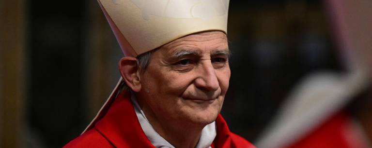 Um cardeal está em destaque, usando vestes religiosas vermelhas e um grande chapéu branco. Ele tem um olhar sereno e está em um ambiente que parece ser uma igreja ou catedral, com outros indivíduos ao fundo. A iluminação é suave, destacando sua expressão facial.