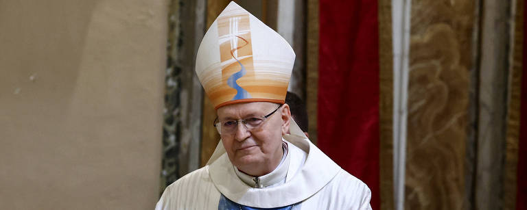 Um homem vestido com vestes litúrgicas brancas e um grande chapéu de bispo, que possui um design em tons de laranja e azul. Ele está em uma posição de oração ou reflexão, com as mãos unidas na frente do corpo. O fundo apresenta uma decoração simples, com cortinas vermelhas e elementos arquitetônicos.