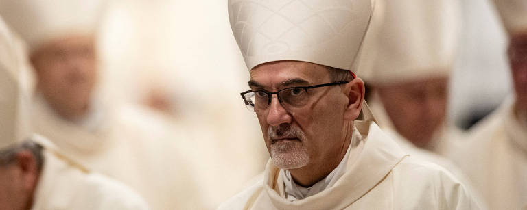 A imagem mostra um homem com um grande chapéu branco, típico de figuras religiosas, em um ambiente de cerimônia. Ele usa óculos e uma vestimenta branca, e parece estar em um momento de reflexão ou oração. Ao fundo, outras pessoas com chapéus semelhantes estão presentes, mas não são claramente visíveis.