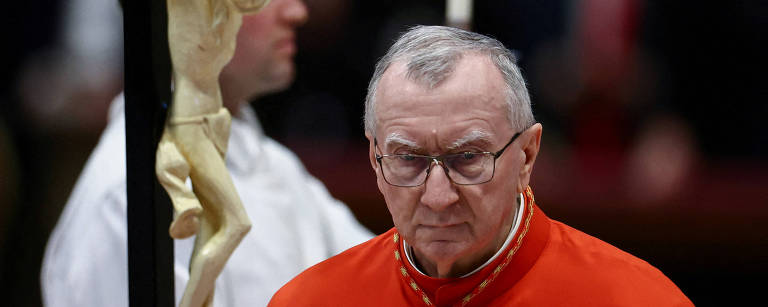 A imagem mostra um homem idoso, vestido com uma vestimenta vermelha, que parece ser um cardeal, com óculos e uma expressão séria. Ao fundo, há uma figura de um crucifixo com a imagem de Jesus crucificado. Um assistente em vestes brancas está ao lado do cardeal, e outras pessoas são visíveis ao fundo, mas não estão claramente focadas.