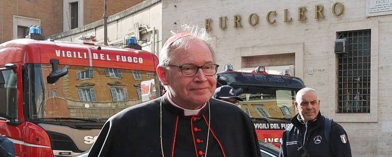 Um cardeal vestido com uma batina preta e um cordão vermelho, carregando uma pasta preta, caminha em uma rua de paralelepípedos em Roma. Ao fundo, há um veículo de emergência e um homem em uniforme de segurança.