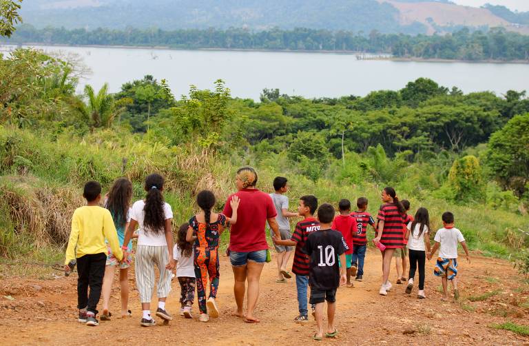 Un gruppo di bambini e adolescenti cammina lungo un sentiero sterrato verso un lago. Si trovano in un ambiente naturale, con vegetazione circostante e la vista del lago sullo sfondo. I bambini sono vestiti in modo casual: alcuni indossano magliette colorate, altri pantaloni o pantaloncini. Il tempo sembra soleggiato e la scena trasmette un senso di gioia e libertà.