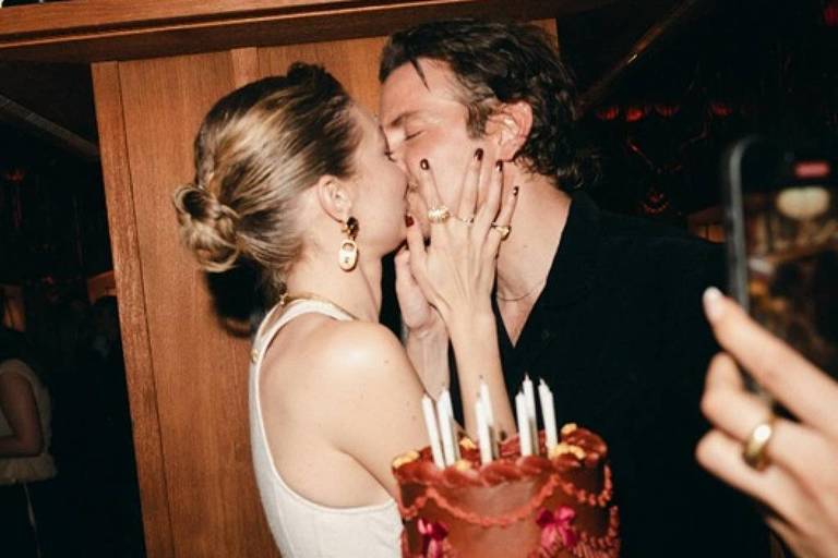 Um casal se beija em um ambiente festivo, com um bolo de aniversário decorado em primeiro plano. O bolo é de chocolate, com estrelas douradas e velas acesas. O fundo mostra prateleiras com livros e outras pessoas ao redor, algumas observando e registrando o momento com celulares. A mulher usa uma blusa branca e brincos, enquanto o homem está vestido de preto.