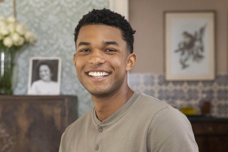 Um homem jovem está sentado em um sofá amarelo, sorrindo para a câmera. Ele usa uma camiseta cinza claro e calças escuras. Ao fundo, há uma mesa de madeira com um vaso de flores brancas e uma parede decorada com papel de parede azul e uma obra de arte. Uma fotografia em preto e branco de uma mulher está visível na parede.
