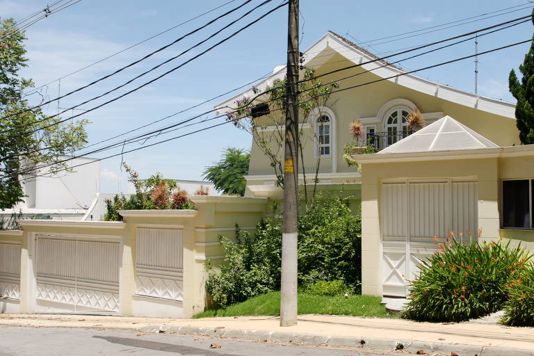 A imagem mostra uma casa de dois andares com uma fachada amarela e um telhado inclinado. Na frente, há um muro com portões brancos e um jardim com plantas verdes. Fios elétricos estão visíveis no céu azul ao fundo, e há árvores ao redor da propriedade.