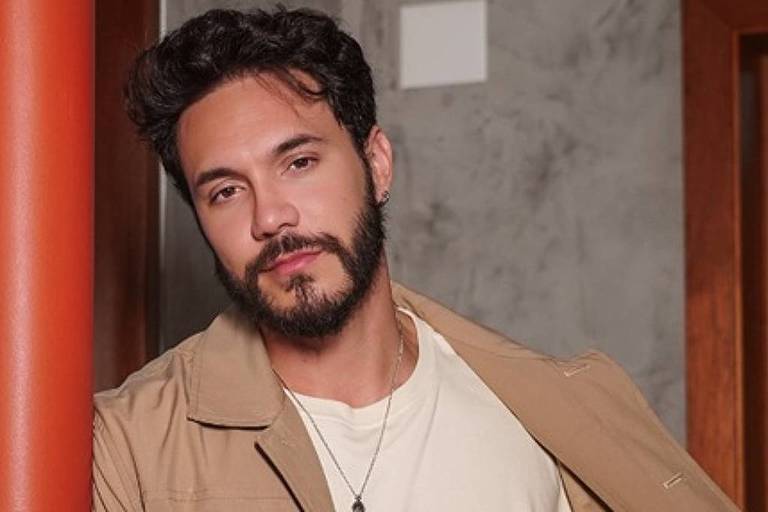 Um homem com barba e cabelo ondulado, vestindo uma jaqueta bege e uma camiseta branca, está posando em um ambiente interno. Ele se apoia em uma coluna laranja e parece olhar para a câmera com uma expressão relaxada. O fundo é composto por paredes de concreto e madeira.