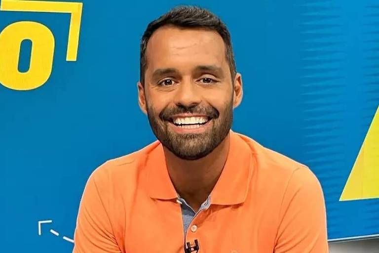 Um homem com cabelo curto e barba bem aparada, vestindo uma camisa polo laranja, está sorrindo para a câmera. O fundo é azul com elementos gráficos em amarelo e branco, possivelmente de um estúdio de televisão.