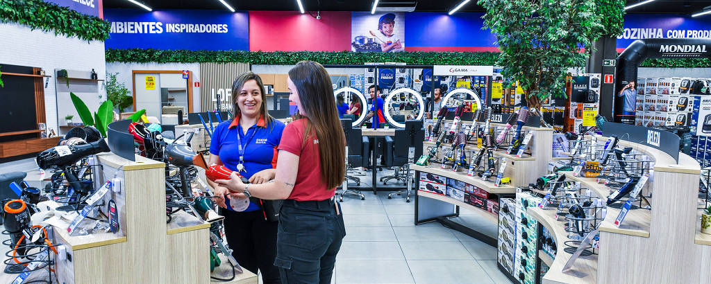A imagem mostra o interior de uma loja de eletrodomésticos, onde duas mulheres estão interagindo. Uma delas, vestindo uma camisa azul, parece ser uma funcionária da loja, enquanto a outra, com uma camiseta vermelha, está segurando um produto. O ambiente é bem iluminado e organizado, com várias prateleiras exibindo eletrodomésticos e utensílios. Ao fundo, há cartazes e plantas decorativas.