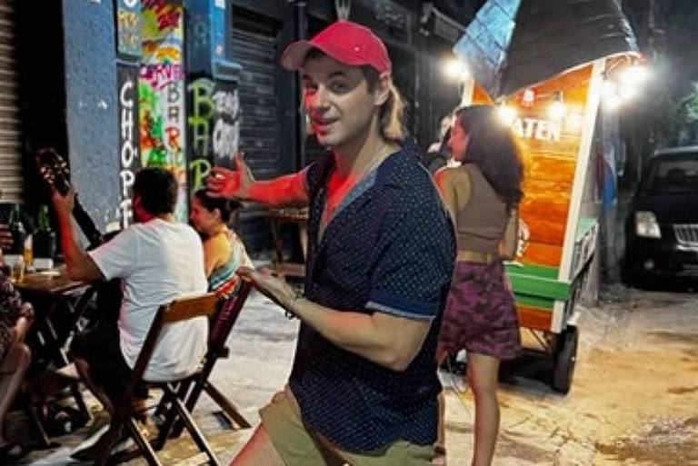 A imagem mostra um homem em uma rua iluminada à noite, usando uma camisa escura e um boné vermelho. Ele está posando com uma perna levantada e um sorriso, enquanto ao fundo há outras pessoas sentadas em mesas, algumas com instrumentos musicais. Um carrinho de comida colorido está visível à direita, e a cena é cercada por paredes grafitadas.