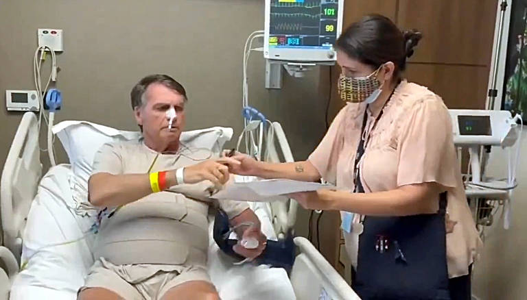 Um homem está sentado em uma cama de hospital, usando uma camisa de hospital e com um tubo de oxigênio no nariz. Ele tem um curativo no braço e está recebendo um documento de uma mulher que está ao seu lado, usando uma máscara. O ambiente é de um quarto de hospital, com equipamentos médicos visíveis ao fundo.