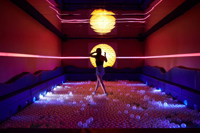A imagem apresenta um ambiente artístico com paredes vermelhas e uma iluminação vibrante. No centro, uma figura humana em silhueta está em uma pose expressiva, cercada por uma superfície coberta de pequenas esferas brancas. Ao fundo, uma grande esfera amarela emite luz, complementada por feixes de luz colorida que atravessam o espaço.