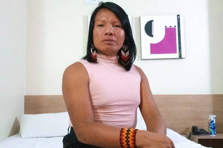 A imagem mostra uma pessoa sentada em uma cama em um ambiente interno. A pessoa está usando uma blusa rosa e uma saia preta, com acessórios de pulseiras laranja e brincos grandes. O fundo é uma parede clara e há uma obra de arte abstrata pendurada. Um objeto azul está visível ao lado da cama.