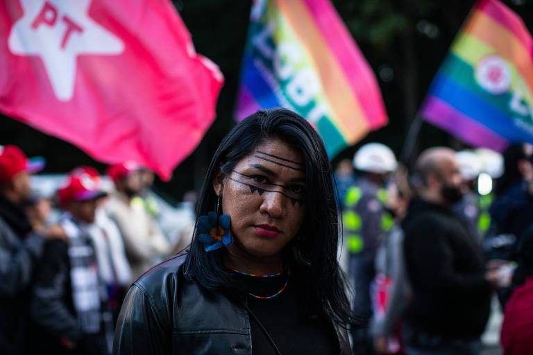 A imagem mostra uma mulher com cabelo longo e escuro, usando uma blusa preta e brincos coloridos. Ela tem marcas de tinta no rosto, com linhas pretas que cruzam suas bochechas. Ao fundo, há várias pessoas e bandeiras coloridas, incluindo bandeiras do movimento LGBTQ+ e uma bandeira com o símbolo do PT. O ambiente parece ser de uma manifestação ou evento público.