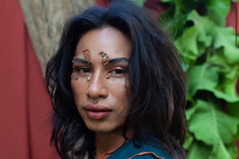 A imagem mostra uma pessoa com cabelo longo e solto, usando uma blusa verde. O rosto está decorado com maquiagem artística, incluindo desenhos que se assemelham a insetos ou elementos naturais. Ao fundo, há uma parede vermelha e folhas verdes, criando um contraste com a figura em primeiro plano.