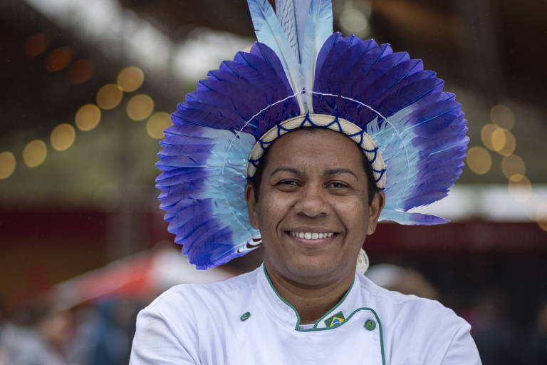 A imagem mostra um chef sorridente, usando um uniforme branco com detalhes verdes e um cocar colorido, predominantemente azul, na cabeça. Ele está posando com os braços cruzados em um ambiente ao ar livre, com pessoas e luzes ao fundo, sugerindo um evento ou festival.