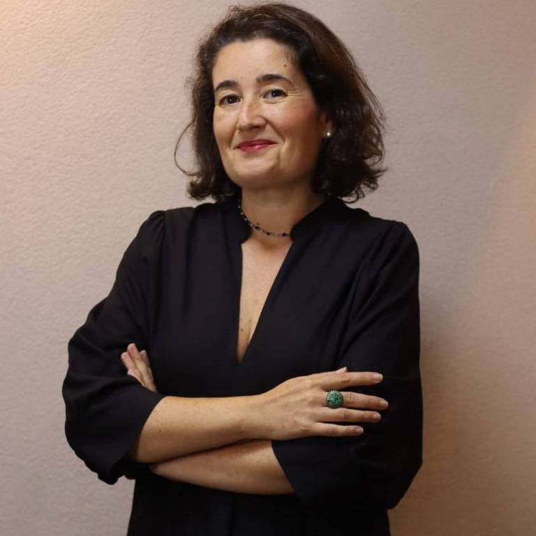 Uma mulher está de pé, com os braços cruzados, usando um vestido preto. Ela tem cabelo castanho escuro e ondulado, e está sorrindo levemente. O fundo é de uma parede clara.