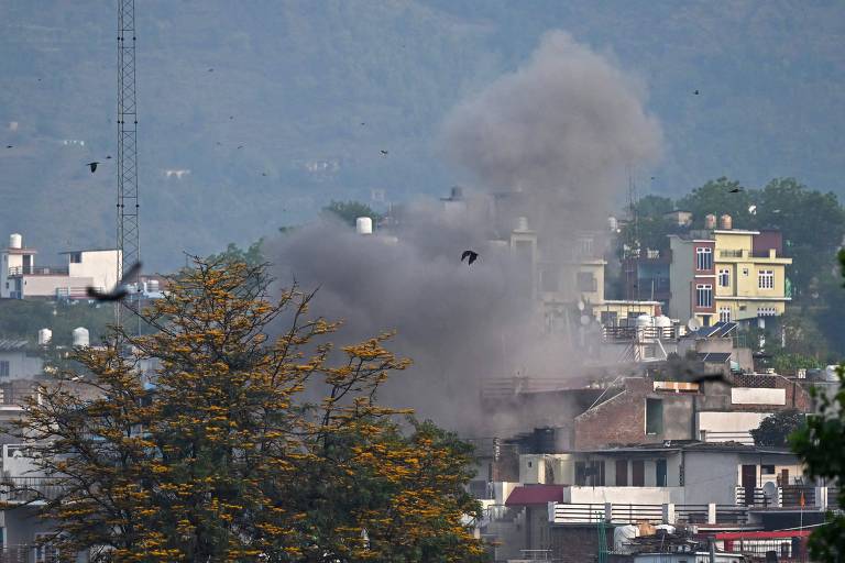 A imagem mostra uma nuvem de fumaça se elevando em uma área urbana, com prédios ao fundo. Há uma árvore com folhas amarelas em primeiro plano e algumas aves voando. O céu parece nublado, e a cena sugere um evento de explosão ou incêndio.