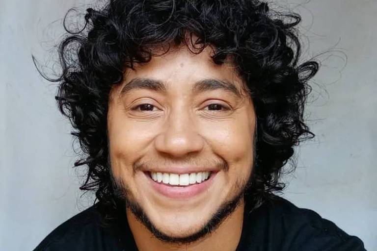 Um homem com cabelo cacheado e barba, sorrindo para a câmera. Ele está vestindo uma camiseta preta e o fundo é neutro, com uma tonalidade clara. O homem parece estar em um ambiente interno, com uma expressão amigável e confiante.