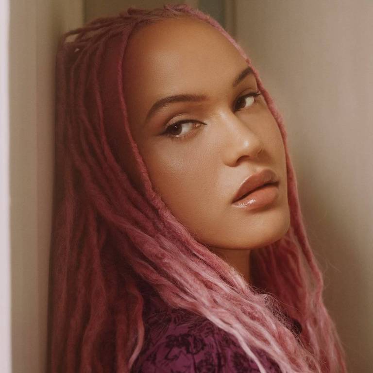 A imagem mostra uma mulher com cabelo longo e dreadlocks em tons de rosa. Ela está encostada em uma parede, olhando para o lado com uma expressão suave. A iluminação é suave, destacando seu rosto e os detalhes do cabelo. A mulher usa um vestido ou blusa de cor escura, que contrasta com o cabelo claro.