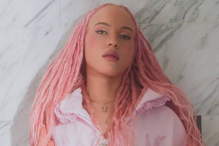 A imagem mostra uma jovem com cabelo rosa e dreadlocks, vestindo um casaco rosa claro com detalhes em renda. Ela está em pé, com uma expressão séria, em frente a uma parede de mármore. O ambiente parece bem iluminado, destacando a textura do casaco e o cabelo da jovem.
