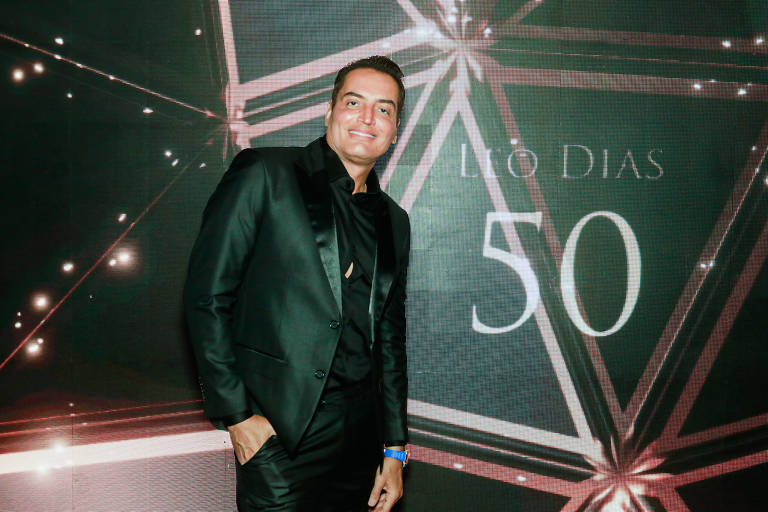 Um homem sorridente está posando em um evento, vestindo um terno preto. Ao fundo, há uma decoração com estrelas e um painel que diz '50' e 'Lúcio Dias'. A iluminação é suave e há um brilho sutil no ambiente.