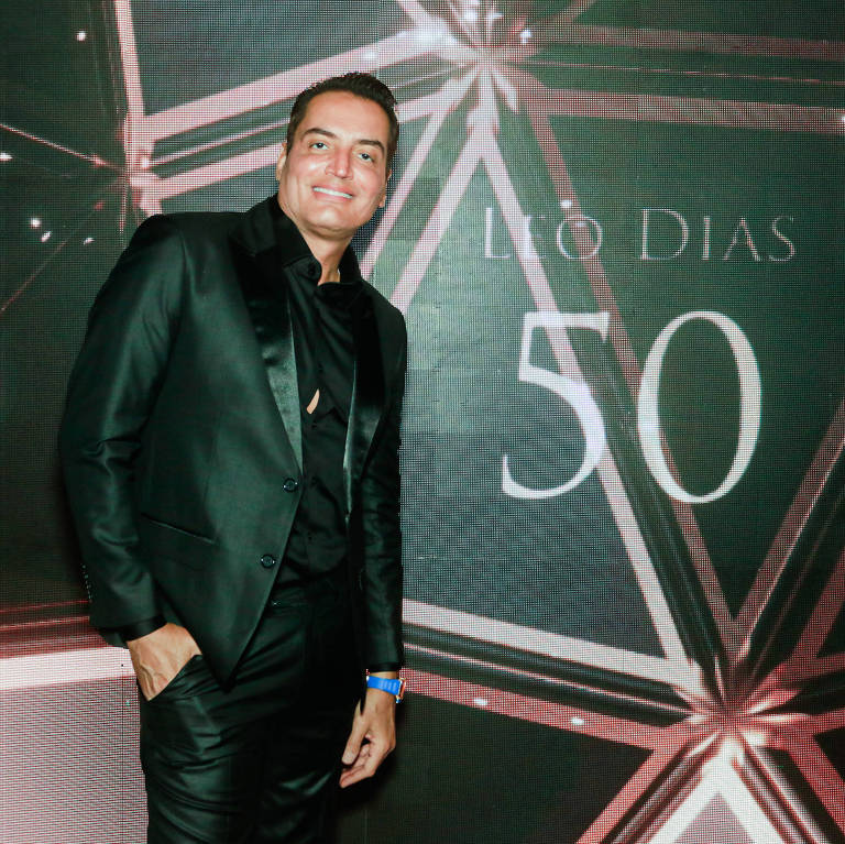 Um homem sorridente está posando em um evento, vestindo um terno preto. Ao fundo, há uma decoração com estrelas e um painel que diz '50' e 'Lúcio Dias'. A iluminação é suave e há um brilho sutil no ambiente.
