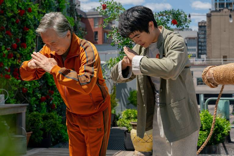 A imagem mostra dois homens em um ambiente externo, possivelmente um jardim ou terraço. O homem à esquerda está vestido com um conjunto de treino laranja e está fazendo uma saudação com as mãos unidas em frente ao peito. O homem à direita, vestido com uma jaqueta clara e camiseta, também está fazendo um gesto semelhante. Ao fundo, há plantas verdes e flores vermelhas, além de edifícios visíveis na cidade.