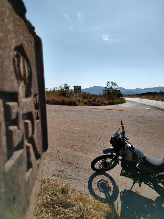 A imagem mostra uma motocicleta estacionada ao lado de uma estrada em um cruzamento. No primeiro plano, há uma placa de sinalização com a letra 'ER' visível. Ao fundo, pode-se ver uma estrada que se bifurca, com montanhas ao longe e um céu claro. A vegetação ao redor é baixa e seca, indicando um ambiente rural.