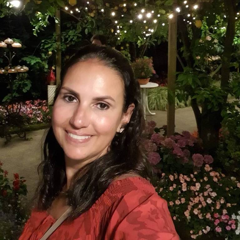 A imagem mostra uma mulher sorrindo em um ambiente ao ar livre, decorado com luzes penduradas e cercado por plantas e flores. O fundo é verde, com uma variedade de plantas e flores coloridas, criando uma atmosfera acolhedora e festiva. A mulher está usando uma blusa vermelha e tem cabelo longo e escuro.