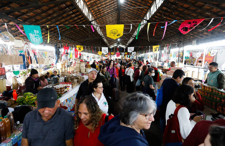 A imagem mostra uma feira interna movimentada, com várias barracas vendendo produtos. Há muitas pessoas circulando entre as barracas, algumas interagindo e outras comprando. O ambiente é iluminado e decorado com bandeirinhas coloridas penduradas no teto. Os produtos expostos incluem frutas, verduras e outros itens típicos de feira.