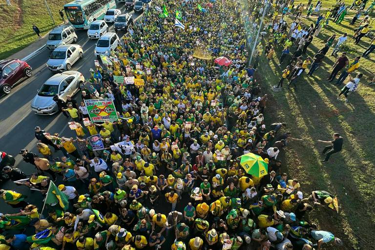 A imagem mostra uma grande multidão de pessoas reunidas em uma manifestação, predominantemente vestidas com camisetas amarelas e verdes, segurando bandeiras do Brasil. A cena é vista de cima, com carros e ônibus parados ao lado da multidão. O ambiente é ao ar livre, com um gramado visível e algumas árvores ao fundo. A atmosfera parece ser de celebração e apoio.