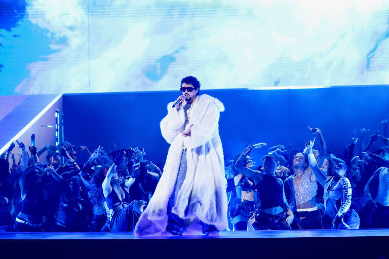 Um artista se apresenta em um palco iluminado com luzes azuis e brancas, vestindo um longo casaco branco. Ele segura um microfone e canta, enquanto um grupo de dançarinos se movimenta ao fundo, criando uma atmosfera vibrante e energética. O cenário é moderno, com projeções visuais ao fundo.