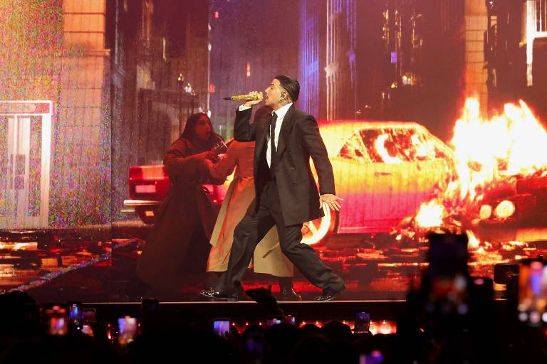 Um artista está se apresentando em um palco, vestido com um terno preto e segurando um microfone. Ao fundo, há uma projeção de uma cena urbana com um carro em chamas e uma figura vestida com um manto. O público está visível na parte inferior da imagem, segurando celulares para gravar a performance.