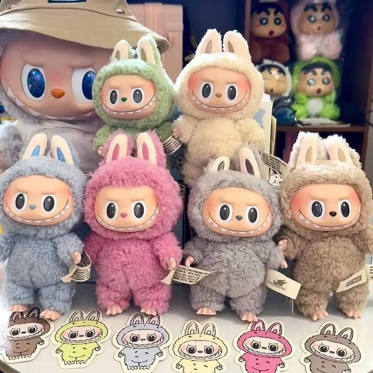 A imagem mostra uma coleção de bonecos de pelúcia em forma de coelho, com diferentes cores como verde, rosa, azul, bege e cinza. Os bonecos têm orelhas de coelho e rostos expressivos. Ao fundo, há um boneco maior com um chapéu e outros bonecos de pelúcia em cores variadas. Na frente, estão adesivos com desenhos de coelhos em diferentes cores.