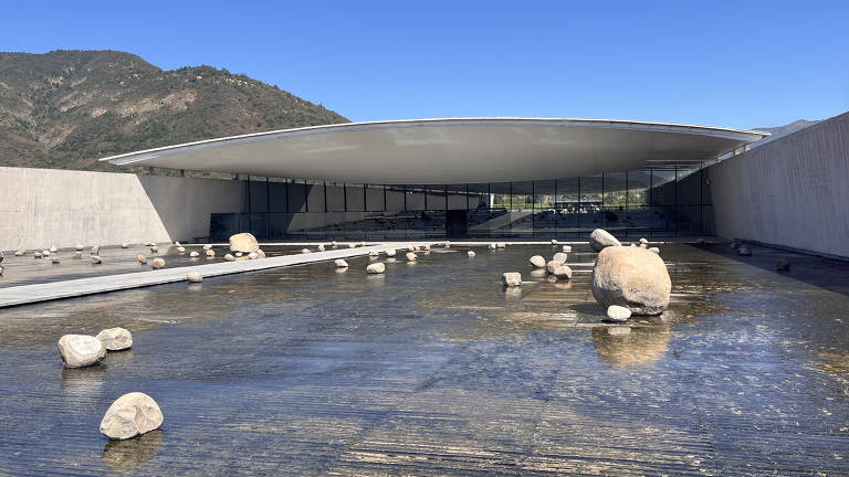 A imagem mostra um edifício moderno com uma estrutura de telhado curvo, feito de concreto e vidro. Na frente do edifício, há um lago raso com várias pedras dispostas de forma irregular. Ao fundo, pode-se ver uma montanha coberta de vegetação sob um céu azul claro.