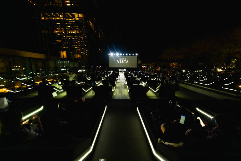 A imagem mostra um cinema ao ar livre à noite, com várias cadeiras dispostas em fileiras. No centro, há um grande telão exibindo a palavra 'CINE VISTA'. As cadeiras têm iluminação em suas bordas, e algumas pessoas estão sentadas, algumas usando dispositivos móveis. Ao fundo, há uma estrutura iluminada que parece ser um edifício.