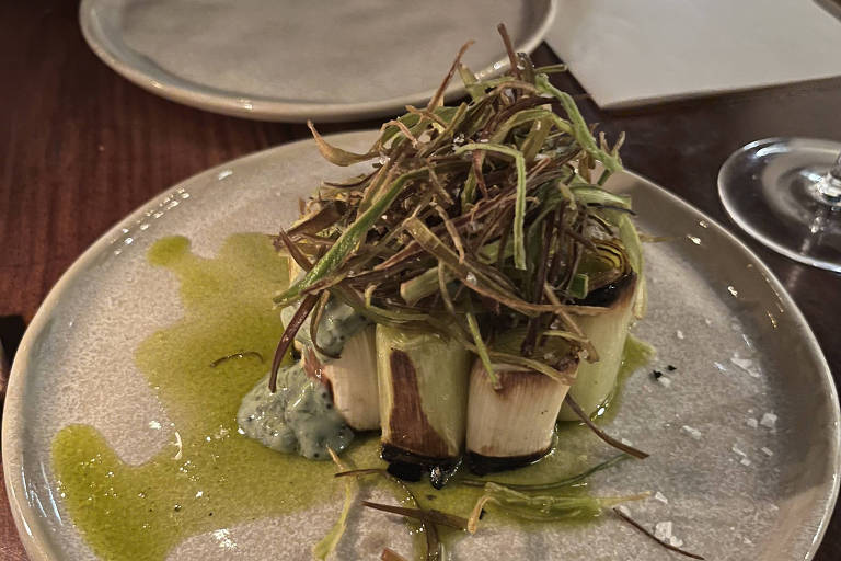 Um prato gourmet apresentado em um prato redondo. No centro, há uma porção de comida que parece ser um tipo de carne ou vegetal, coberta com uma camada de ervas finas. Ao redor, há um molho verde e algumas gotas de um líquido claro. O fundo é de madeira escura, e há um copo de vinho ao lado.