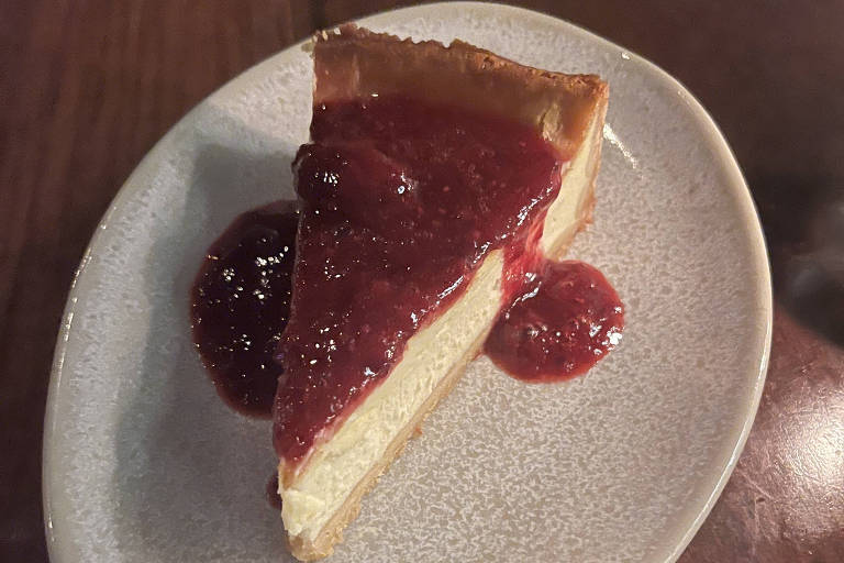 Uma fatia de cheesecake é apresentada em um prato oval. A torta é coberta com uma camada de compota de frutas vermelhas, que se espalha sobre a superfície do cheesecake. O prato é de cor clara e a mesa ao fundo é de madeira escura.