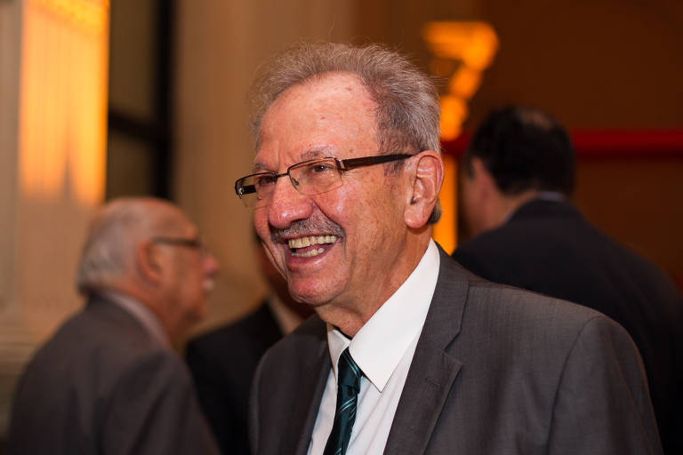 Um homem de cabelos grisalhos e bigode, usando óculos e um terno cinza, sorri enquanto conversa em um evento. Ao fundo, outras pessoas em trajes formais estão visíveis, mas não são claramente identificáveis. A iluminação do ambiente é suave, com um tom quente.