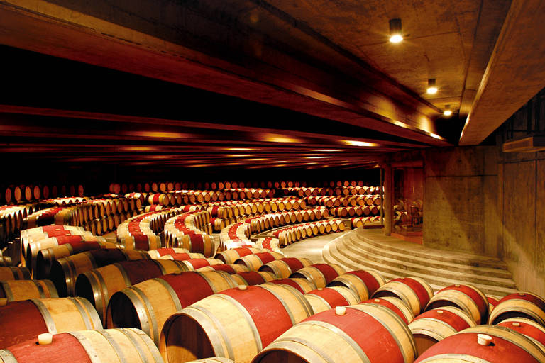 A imagem mostra o interior de uma adega, onde barris de vinho esto organizados em fileiras. Os barris so de madeira, com a parte superior em tom claro e a parte inferior em tom escuro, formando um padro. A iluminao  suave, destacando a textura dos barris e as paredes de concreto da adega. O ambiente  amplo e bem estruturado, com um piso em espiral que se destaca na composio.