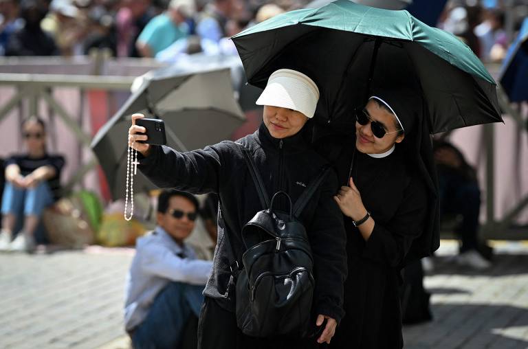 Duas mulheres, uma vestida de freira e outra com um chapéu branco, estão tirando uma selfie em um local ao ar livre. A mulher de chapéu está segurando um celular e um terço, enquanto a freira está ao seu lado, usando óculos escuros e segurando um guarda-chuva. Ao fundo, há várias pessoas sentadas e um ambiente movimentado.