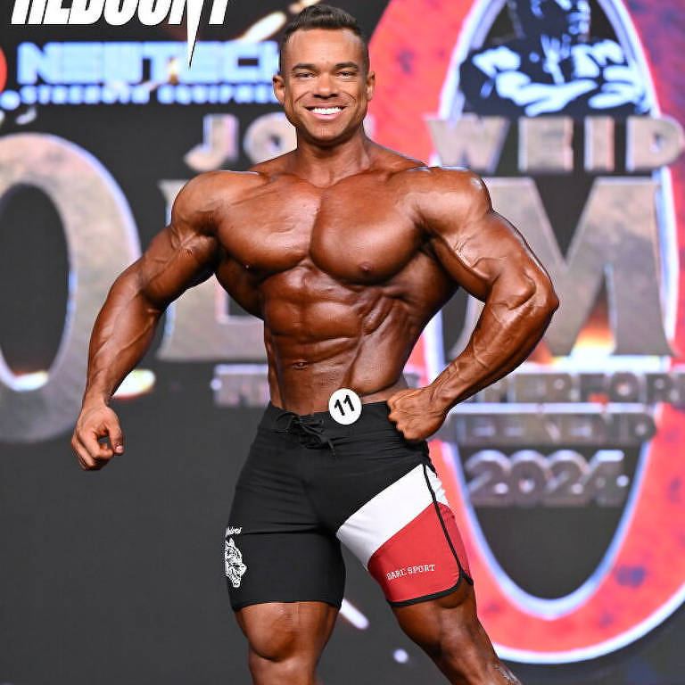 Um competidor de fisiculturismo está posando no palco durante uma competição. Ele tem um corpo musculoso e bem definido, com destaque para os músculos do peito, braços e pernas. O competidor usa um short de competição preto e vermelho, e está sorrindo enquanto exibe sua forma física. Ao fundo, há um banner com o logotipo da competição e outros elementos gráficos relacionados ao evento.