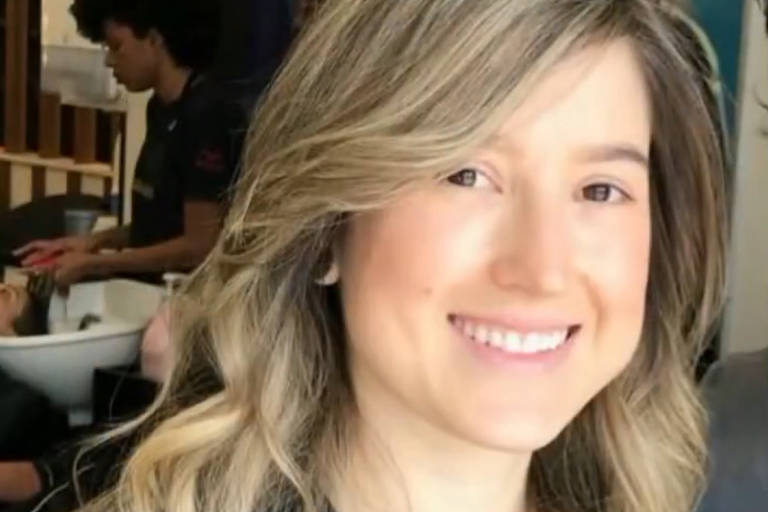 A imagem mostra uma mulher sorridente com cabelo longo e ondulado, em um salão de beleza. Ao fundo, é possível ver outra pessoa em uma cadeira de cabeleireiro, recebendo um tratamento de cabelo. O ambiente é bem iluminado e decorado de forma moderna.