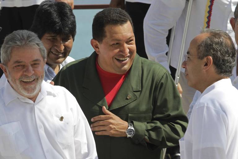 A imagem mostra quatro homens sorrindo e conversando em um evento ao ar livre. Um deles, à esquerda, usa uma camisa branca e tem barba. O segundo, ao centro, está vestido com um uniforme verde e uma camiseta vermelha, sorrindo amplamente. O terceiro, atrás, tem cabelo escuro e está sorrindo. O quarto homem, à direita, usa uma camisa branca e óculos. O fundo é um céu claro e água.