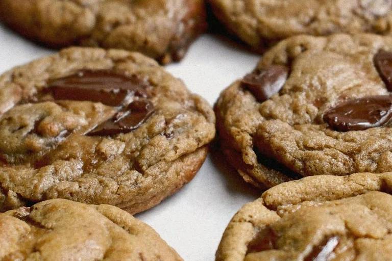 A imagem mostra uma variedade de biscoitos de chocolate dispostos em uma superfície clara. Os biscoitos têm uma textura macia e estão decorados com pedaços de chocolate visíveis em sua superfície. Eles são de cor marrom claro a escuro, com um aspecto caseiro e apetitoso.